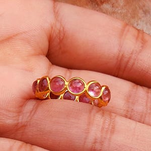Könnte beinhalten: Ein goldener Ring mit einer Reihe von rosa Edelsteinen. Der Ring hat ein goldenes Band und eine Reihe von oval geformten rosa Steinen. Der Ring wird auf der Handfläche einer Person gezeigt, mit einem Holzuntergrund im Hintergrund.