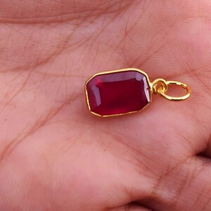 Könnte beinhalten: Ein rechteckiger Anhänger mit tiefrotem Edelstein, goldfarbener Lünette und Schlaufe. Der Anhänger wird in einer Hand gehalten, wodurch die satte Farbe und das Design des Schmuckstücks zur Geltung kommen. Die Goldeinfassung verstärkt den rubinartigen Stein.