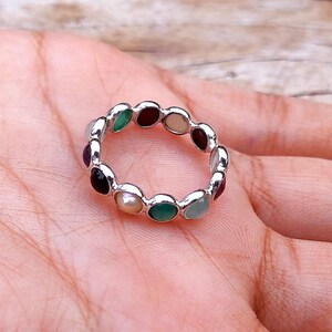 Puede incluir: Un anillo de plata con una serie de piedras preciosas de colores. El anillo presenta una variedad de piedras, incluyendo verdes, rojas, negras y blancas, dispuestas en un patrón circular. El anillo se muestra en la mano de una persona.