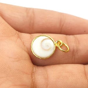K&ouml;nnte beinhalten: Ein goldfarbener Anh&auml;nger mit einem wei&szlig;en, spiralf&ouml;rmigen Design. Der Anh&auml;nger ist rund und hat einen goldenen Rand. Er wird in der Hand einer Person gehalten.