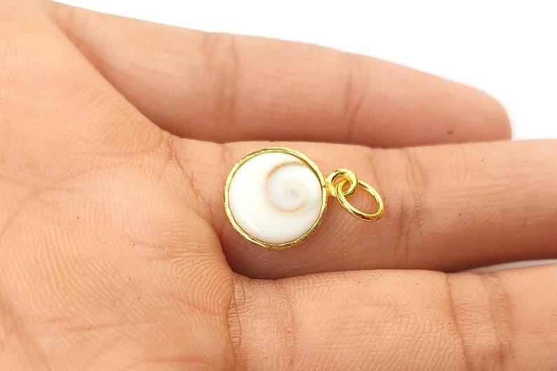 K&ouml;nnte beinhalten: Ein goldfarbener Anh&auml;nger mit einem wei&szlig;en, spiralf&ouml;rmigen Design. Der Anh&auml;nger ist rund und hat einen goldenen Rand. Er wird in der Hand einer Person gehalten.