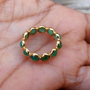 May include: A gold-colored ring featuring a continuous band of oval-shaped, vibrant green gemstones. The ring is set against a skin-toned background, highlighting the contrast between the gold and green elements. The ring's design suggests a classic, elegant style.
