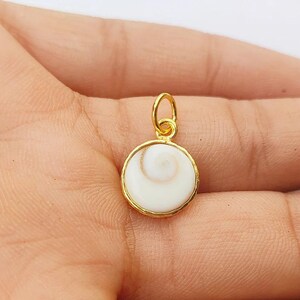 K&ouml;nnte beinhalten: Ein goldfarbener Anh&auml;nger mit einem wei&szlig;en, spiralf&ouml;rmigen Design. Der runde Anh&auml;nger hat einen goldfarbenen Rand und eine kleine Schlaufe f&uuml;r eine Kette. Der Anh&auml;nger ist wahrscheinlich aus Metall und einem wei&szlig;en Stein gefertigt.