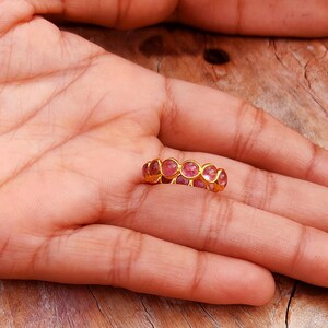 May include: A gold ring with a series of pink gemstones. The ring is set in a gold band and features a row of oval-shaped pink stones. The ring is displayed on a person's palm, with a wooden surface in the background.