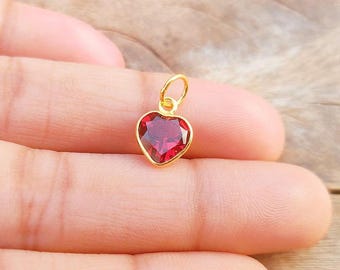 May include: A gold-coloured heart-shaped pendant with a vibrant red gemstone. The pendant is suspended from a small loop, and the heart's edges are smooth and rounded. The gemstone is faceted, catching the light.