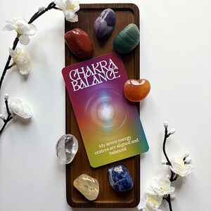 May include: A rectangular wooden tray holds a Chakra Balance card with a gradient background and the text My seven energy centres are aligned and balanced. Seven polished, multi-coloured stones are arranged around the card on the tray.