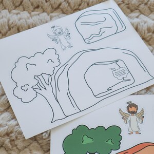 May include: White paper cutouts featuring line drawings of an angel, a tree, a cave, and a rock. One cutout has a speech bubble with text. The angel has a halo and wings. The tree is green. The cave is white with black outlines.
