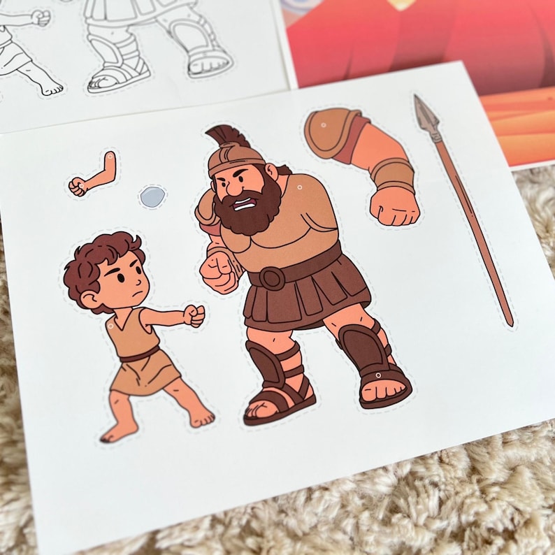 May include: A set of illustrated paper cutouts depicting a biblical scene. The main figures are a large warrior in brown armor and a smaller boy in a tunic. A spear and additional cutout pieces are also visible.