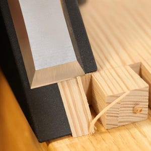 Könnte beinhalten: Nahaufnahme der Holzbearbeitung. Ein Metallmeißel mit schwarzem Griff schneidet in einen hellen Holzblock. Holzspäne sind sichtbar, was den Prozess der Holzformung anzeigt. Die Szene ist gut beleuchtet und hebt die Details des Handwerks hervor.