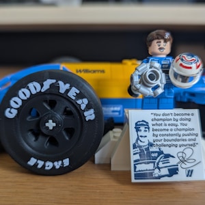 May include: A blue and yellow Lego Formula 1 race car with a Goodyear tire, a Lego driver figure holding a helmet, and a sticker with a quote about becoming a champion.