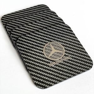 May include: A set of four black carbon fiber coasters with a silver Mercedes-Benz logo and the text "Mercedes-Benz".
