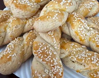 May include: A plate of baked, twisted breadsticks, sprinkled with sesame seeds. The breadsticks have a golden-brown glaze and are arranged on a white plate. The baked goods are a light tan color.