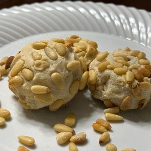 May include: Two round, pale-colored cookies covered in light brown pine nuts. The cookies are resting on a white plate with a ridged, scalloped edge. Several loose pine nuts are scattered across the surface of the plate around the cookies.