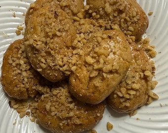 May include: A plate of golden-brown cookies, oval-shaped and topped with chopped nuts. The cookies are arranged in a cluster, with some nuts scattered around them. The plate is white with a ridged design.