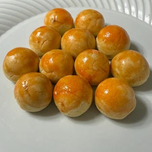 May include: A white plate holds a dozen golden-brown, round pastries. Each pastry has a glossy, orange-tinted top. The baked goods are arranged in a neat, slightly overlapping pattern, suggesting a dessert or snack.