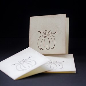 May include: Three blank cards with a brown outline of a pumpkin on the front. The cards are off-white and have a slightly textured surface.