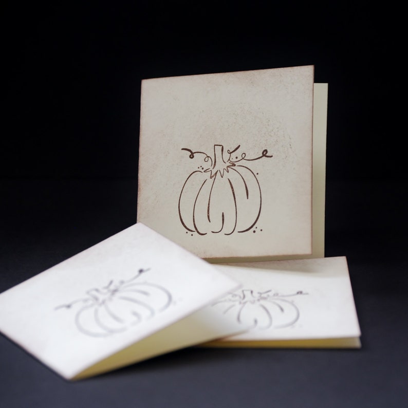 May include: Three blank cards with a brown outline of a pumpkin on the front. The cards are off-white and have a slightly textured surface.