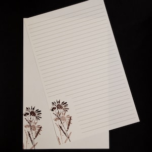 May include: Cream-colored notepad with horizontal lines for writing. The bottom left corner features two brown floral designs. The notepad is open, revealing the lined pages, and is set against a black background.
