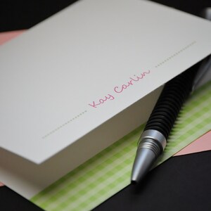 May include: A white card with a green and white checkered pattern on the bottom. The card has a pink inscription that says "Kay Carlin" with a dotted line above and below the name.