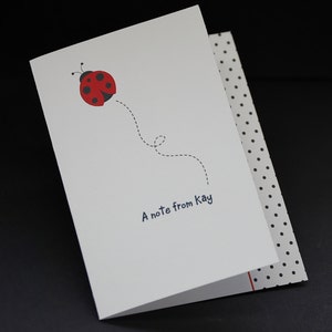 Peut inclure: Une carte blanche avec une coccinelle rouge à points noirs volant avec une ligne pointillée. La carte dit "A note from Kay" en bas.