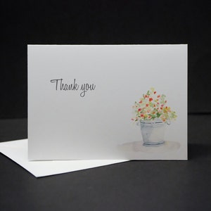 May include: A white card with the text "Thank you" printed in black. A watercolour illustration of a white pot with red, yellow, and white flowers is on the right side of the card.