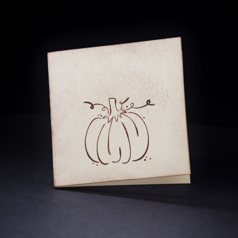 May include: A blank card with a brown line drawing of a pumpkin on it. The card is off-white and has a slightly textured surface.