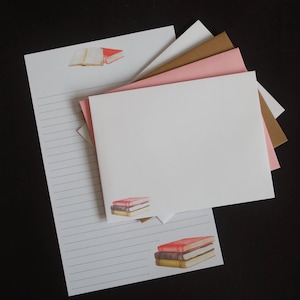 May include: A set of blank stationery featuring a stack of books with a red, brown, and yellow cover. The stationery is white with lined paper and comes with envelopes in pink, brown, and white.
