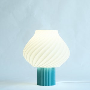 May include: A modern table lamp with a white, ribbed lampshade and a teal base. The lampshade has a soft, curved shape, and the base is cylindrical. The lamp is plugged into a white cord.