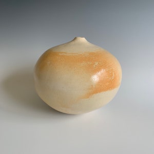 May include: A white ceramic vase with a rounded shape and a subtle orange glaze. The vase has a smooth, glossy finish and a small opening at the top.