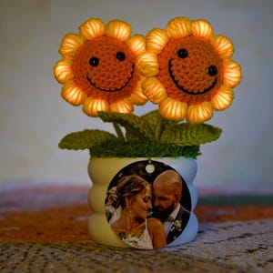 May include: A decorative potted plant featuring two crocheted sunflowers with smiling faces. The sunflowers are orange and yellow, with green leaves and a white pot. A circular tag with a photo of a couple is attached to the pot.