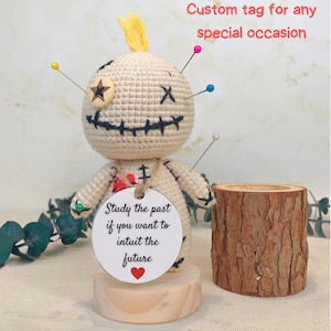May include: A handmade beige crochet voodoo doll with pins, a wooden star, and a stitched mouth. It holds a tag that reads "Study the past if you want to intuit the future." The doll is displayed with a wooden candle holder. Text above reads "Custom tag for any special occasion."