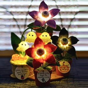 May include: A collection of handmade crochet Halloween decorations. The arrangement includes illuminated flowers in purple, red, and black, along with yellow ghost figures. Each piece is potted and adorned with festive tags. The text on the tags reads: "Witching you a spook-tacular Halloween!" and "A candy a day keeps the monsters away."