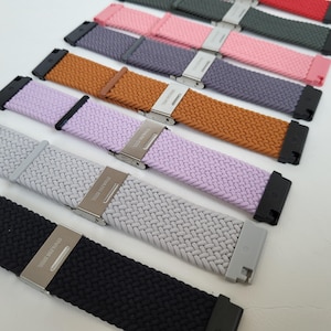 Puede incluir: Un conjunto de ocho correas de reloj tejidas en varios colores, incluyendo negro, gris, morado claro, naranja, rosa, rojo y azul. Las correas están hechas de un material suave y flexible y cuentan con una hebilla plateada.