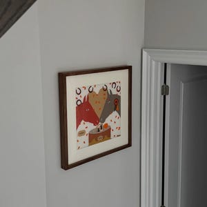 May include: A framed art print featuring three horses gathered around a birthday cake. The horses are depicted in red, tan, and gray, with confetti and horseshoe accents. The artwork is set against a white background and framed in a dark wood.