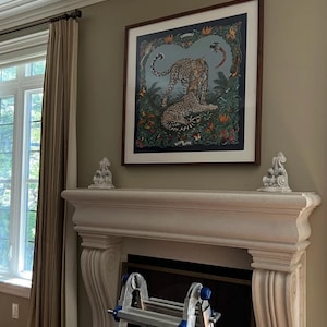 May include: A framed art print featuring two leopards in a jungle setting. The artwork is in a dark wooden frame and hangs above a white fireplace mantel. The print has a blue background with colorful flora and fauna.