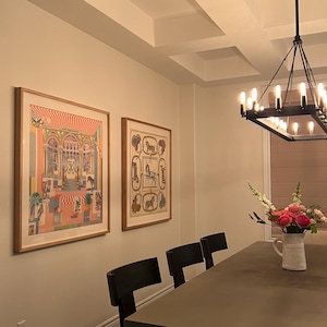 May include: Two framed rectangular wall art prints featuring intricate, colorful architectural and equestrian illustrations, displayed side-by-side on a light-colored wall above a dining table with black chairs and a white floral pitcher.