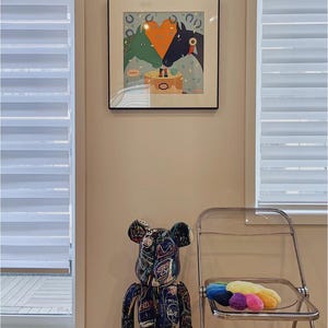 May include: A black and white graffiti-style Bearbrick toy stands next to a clear plastic chair with a rainbow-coloured plush toy on the seat. A framed print of two horses with a heart-shaped orange object between them hangs on the wall.