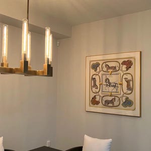 May include: A framed artwork featuring a detailed illustration of horses and equestrian-themed elements in a cream-colored frame. The artwork is displayed on a light gray wall, with a modern chandelier and dining table visible.