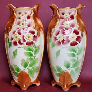 May include: Pair of decorative ceramic vases. Each vase features a cream-colored body with a floral design in shades of red, pink, and yellow. Green leaves and brown accents frame the floral arrangement. The vases have a unique shape and are supported by three small feet.