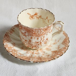 Peut inclure: Un service à thé vintage composé d'une tasse et d'une soucoupe. La tasse et la soucoupe sont blanches avec un motif floral détaillé dans des tons orange et marron. Les bords sont festonnés et dorés.