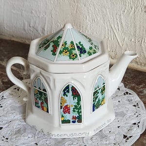 Puede incluir: Una tetera de cerámica blanca con forma de invernadero y tapa hexagonal. El cuerpo presenta ilustraciones de ventanas arqueadas con plantas y flores coloridas. El texto 'a conservatory teapot' está impreso en negro a lo largo de la base.