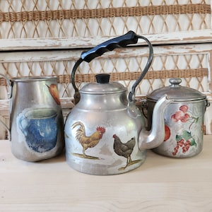 May include: Three vintage aluminum teapots with decorative designs. The teapot in the center has a rooster and a hen design. The teapot on the left has a blue cup design. The teapot on the right has a red berry design.