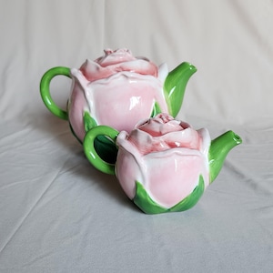 May include: Two ceramic teapots shaped like blooming pink roses with green leaves at the base, green handles, and green spouts. Each teapot features a lid designed as the centre of the rose flower, with a glossy finish and soft pink petals.