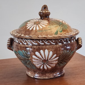 May include: Brown ceramic lidded serving bowl with white daisy and green leaf designs. The bowl has two handles and a decorative finial on the lid. The rim of the lid has a white and brown pattern.