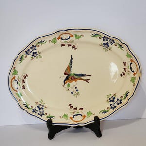 Peut inclure: Un grand plat de service ovale en céramique avec un fond crème et un motif d'oiseau bleu, vert et orange. Le plat a un bord festonné et est décoré de motifs floraux.