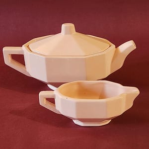 Puede incluir: Un juego de tetera y jarra de crema de cerámica rosa con un diseño geométrico. La tetera tiene una tapa y un pico. La jarra de crema tiene un asa y un pico.