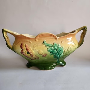 Peut inclure: Un cache-pot en céramique à deux anses, présentant un dégradé de couleurs allant d'une base vert foncé à un sommet jaune clair. L'extérieur est orné de motifs floraux en relief, incluant une fleur proéminente et des feuilles vertes.