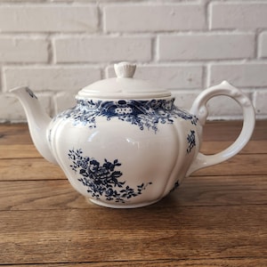 Peut inclure: Une théière en céramique blanche ornée d'un motif floral bleu foncé et de volutes complexes. La théière présente un corps arrondi et côtelé, une anse incurvée, un couvercle assorti avec un petit bouton et un bec verseur à motifs bleus.