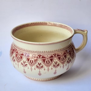 Può includere: Un vaso da notte in ceramica bianca con un motivo floreale rosso e bianco. Il vaso ha un'unica maniglia e un corpo arrotondato.