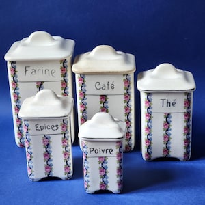 Può includere: Cinque barattoli di spezie in ceramica bianca con coperchi, ognuno etichettato con una parola francese diversa. I barattoli sono decorati con strisce verticali di motivi floreali rosa e blu. Le parole sui barattoli sono "Farine", "Café", "Épices", "Poivre" e "Thé".